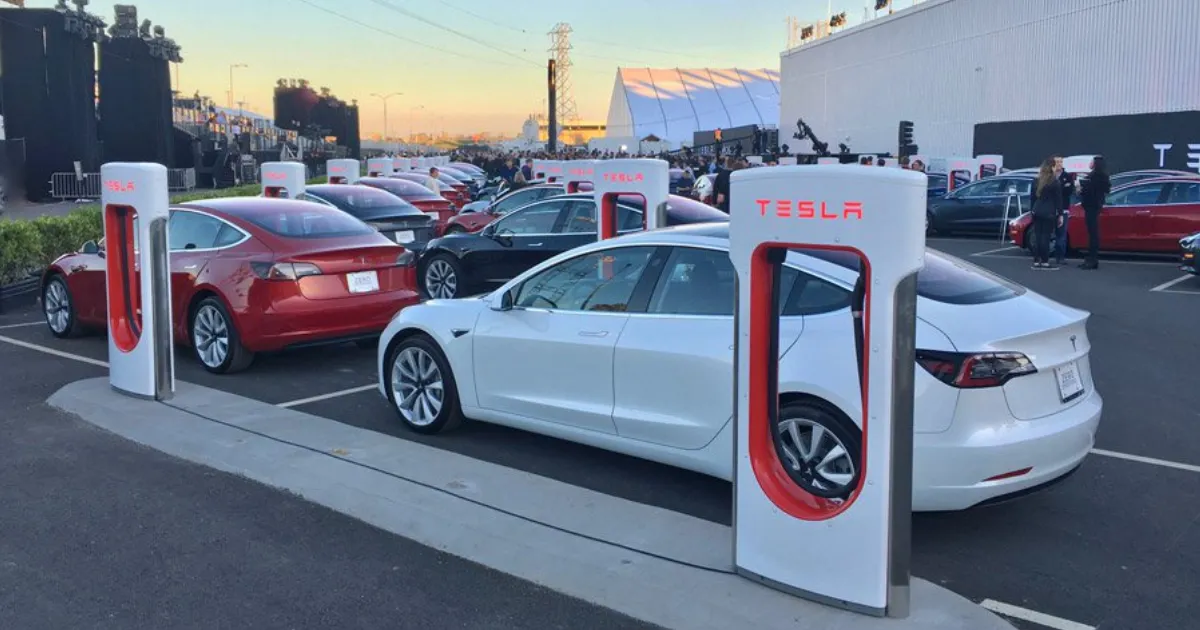 Tesla cars charging at a Tesla Supercharger station in a parking lot, with people and a large building in the background.