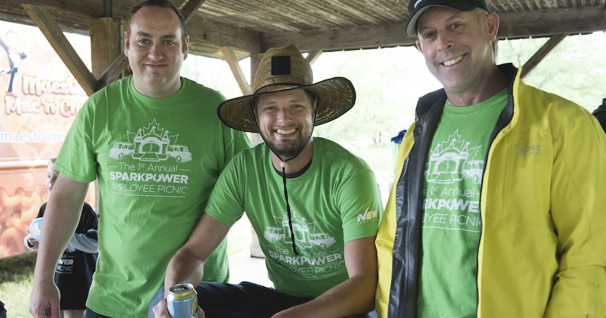 Three men in matching green shirts at a picnic, one wearing a straw hat, smiling under a wooden shelter.