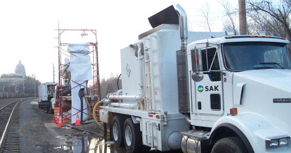 A SAK construction truck is parked beside railway tracks, with scaffolding and orange cones nearby. A distant building is in the background.