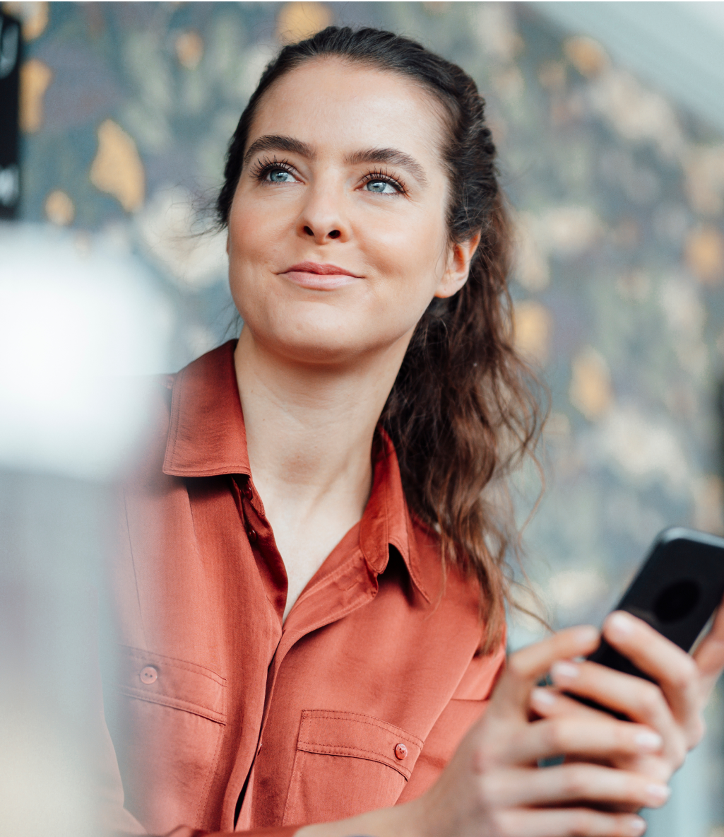 Woman in a red shirt holding a smartphone, looking to the side, with a blurred background featuring floral patterns.