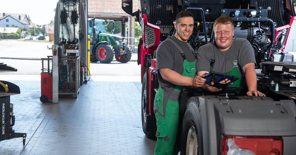 Two mechanics in overalls stand smiling beside a truck in a garage, holding tools. A tractor is visible outside in the background.