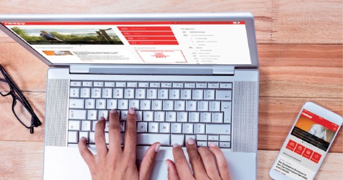 Hands typing on a laptop with a website on screen; a smartphone displaying a similar site is placed nearby on a wooden surface.